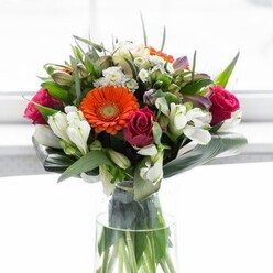 A vision of refined beauty, this bouquet channels the artistry of Tufnell Park's florists, melding colour and form into a harmonious display. Four exquisite gerberas-two glowing orange, two pale peach-anchor the composition, their plush petals radiant in the soft light filtering through a sunlit window, much like the peaceful interiors of homes along Campdale Road. Clustered among them are magenta roses, each bloom tasseled and full, suggestive of romance and grace found in the area's quiet garden squares. Sprays of pure white alstroemeria peek from amidst the fuller blossoms, lending gentle brightness and a whisper of fragrance that recalls cool spring mornings in Tufnell Park. The bouquet's structure is defined by broad, enveloping emerald leaves, artfully folded in a protective circle at the base, while finer greenery weaves upward, creating dynamic movement and depth. Arranged in a clear, slender vase, every detail from glossy green stems to subtle shadows can be admired, resulting in a display of balance, freshness, and subtle opulence. This arrangement radiates warmth and hope-perfect as a centrepiece for a gathering of friends, a special gift for a milestone, or a thoughtful gesture of appreciation. The intricate layering and rich palette conjure memories of colourful market stalls and lush local gardens, delivering a taste of Tufnell Park's vibrant character to any occasion.
